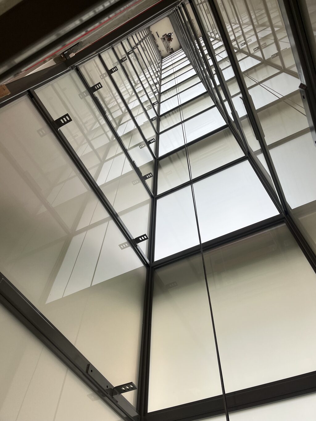 Blick von unten durch einen verglasten Aufzugsschacht aus Stahl